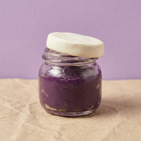 A clay jar of purple ube jam with a cream lid, against a lilac wall